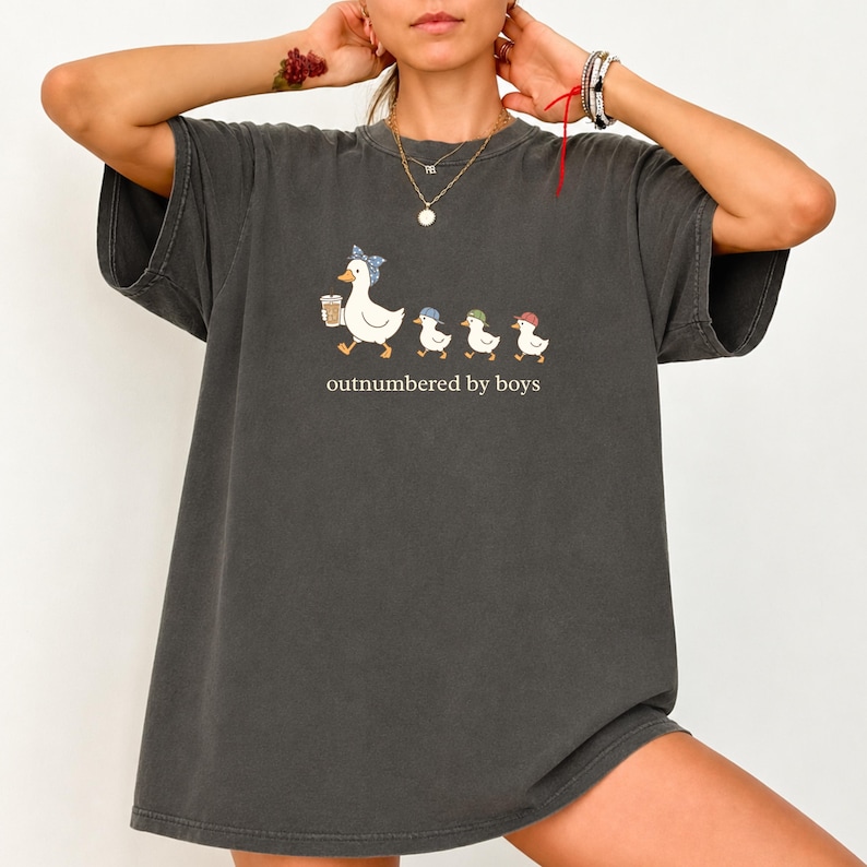 May include: A charcoal gray short-sleeve t-shirt featuring a graphic of a mother duck wearing a blue polka dot headscarf holding a coffee cup, followed by three small ducklings in colorful baseball caps, with the text outnumbered by boys.