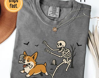 May include: A charcoal gray crew neck t-shirt featuring a graphic print of a skeleton chasing a corgi dog holding a bone in its mouth. The scene includes small bats, a bare tree, and a tombstone. The shirt has a Comfort Colors brand label.