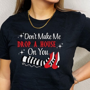 Op de afbeelding: Zwart T-shirt met de tekst "Don't Make Me Drop A House On You" en een afbeelding van een paar rode schoenen met zwart-wit gestreepte benen.