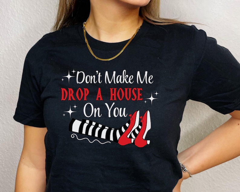 Op de afbeelding: Zwart T-shirt met de tekst "Don't Make Me Drop A House On You" en een afbeelding van een paar rode schoenen met zwart-wit gestreepte benen.