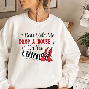 Op de afbeelding: Witte trui met een zwart en rood motief van heksenbenen en schoenen. De tekst op de trui luidt "Don't Make Me Drop A House On You".