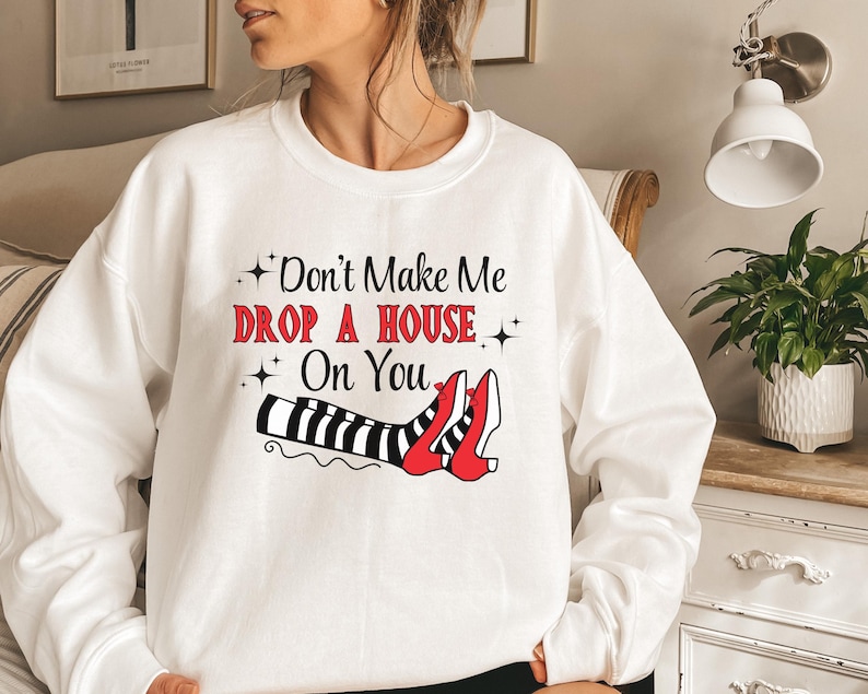 Op de afbeelding: Witte trui met een zwart en rood motief van heksenbenen en schoenen. De tekst op de trui luidt "Don't Make Me Drop A House On You".