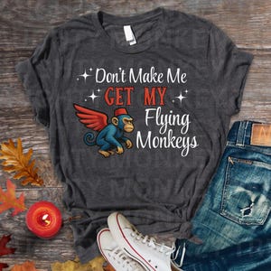May include: Dark gray t-shirt with the text "Don't Make Me Get My Flying Monkeys." The shirt features a cartoon monkey with red wings and a fez. The shirt is paired with blue jeans and white sneakers.