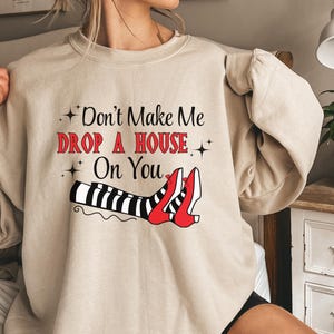 Op de afbeelding: Een beige sweatshirt met een afbeelding van heksenbenen met rode schoenen en zwart-wit gestreepte kousen. De tekst op het sweatshirt luidt "Don't Make Me Drop A House On You".