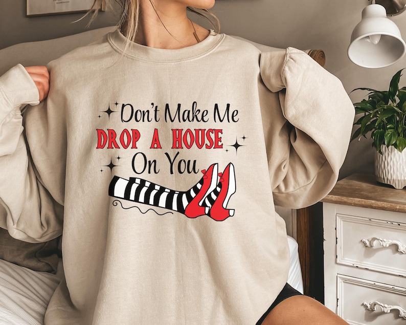 Op de afbeelding: Een beige sweatshirt met een afbeelding van heksenbenen met rode schoenen en zwart-wit gestreepte kousen. De tekst op het sweatshirt luidt "Don't Make Me Drop A House On You".
