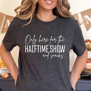 May include: A dark gray t-shirt with the text "Only here for the halftime show and snacks" in white.
