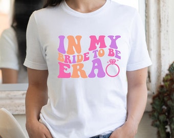May include: White t-shirt with the text "In My Bride To Be Era" in pink, orange, and purple retro font. A pink diamond ring with sparkles is next to the text.