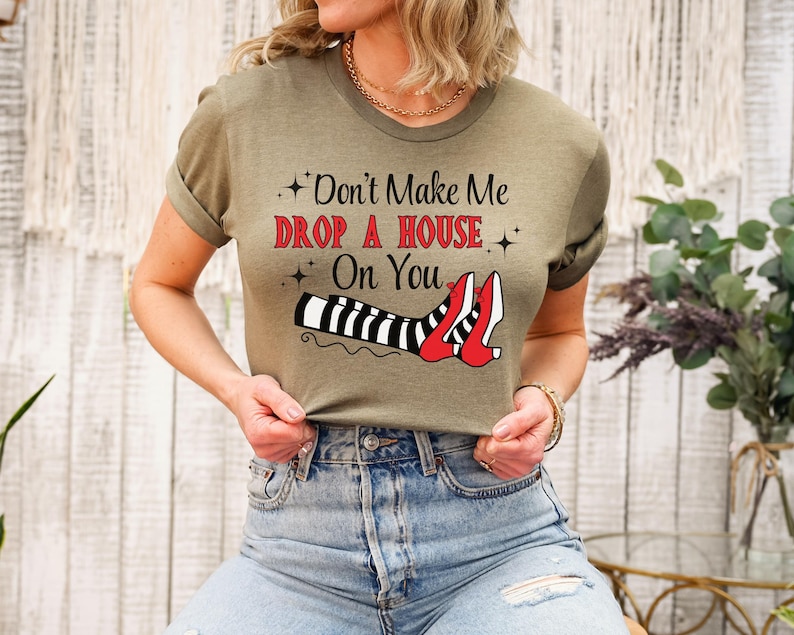 Op de afbeelding: Een vrouw draagt een groen T-shirt met de tekst "Don't Make Me Drop A House On You" en een afbeelding van heksenbenen met rode schoenen en zwart-wit gestreepte kousen.