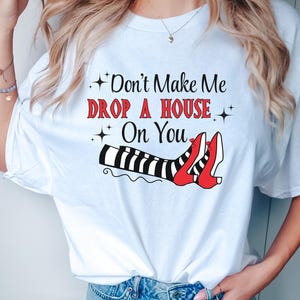Op de afbeelding: Wit T-shirt met een afbeelding van heksenbenen met rode schoenen en de tekst "Maak me niet een huis op je laten vallen".