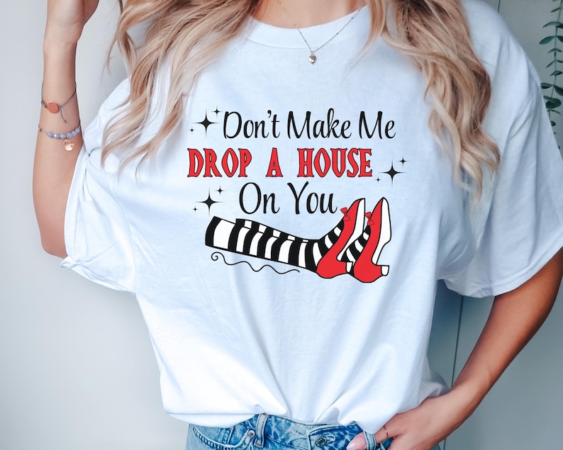 Op de afbeelding: Wit T-shirt met een afbeelding van heksenbenen met rode schoenen en de tekst "Maak me niet een huis op je laten vallen".