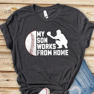 May include: A dark grey t-shirt with a white baseball graphic and the text "My son works from home".