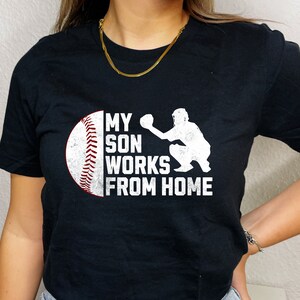 May include: Black t-shirt with a white baseball and catcher graphic. The text on the shirt reads "My son works from home."