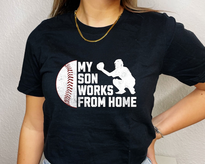 May include: Black t-shirt with a white baseball and catcher graphic. The text on the shirt reads "My son works from home."