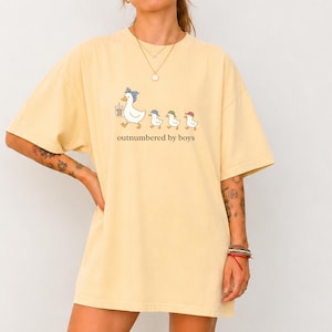 May include: A pale yellow short-sleeve t-shirt featuring a graphic of a mother duck wearing a blue bandana and holding a drink, followed by three small ducklings in colorful caps, with the text "outnumbered by boys" printed below.