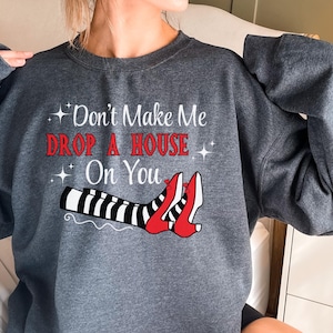 Op de afbeelding: Een donkergrijs sweatshirt met de tekst "Don't Make Me Drop A House On You" en een afbeelding van een paar rode schoenen met zwart-wit gestreepte benen.