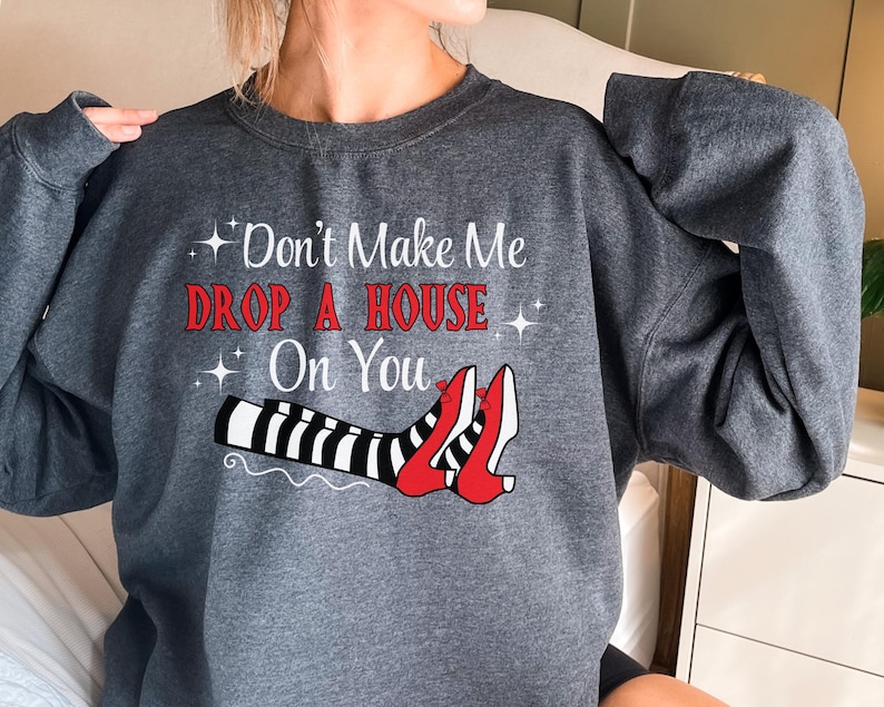 Op de afbeelding: Een donkergrijs sweatshirt met de tekst "Don't Make Me Drop A House On You" en een afbeelding van een paar rode schoenen met zwart-wit gestreepte benen.