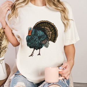 May include: A white t-shirt with a colorful illustration of a turkey. The turkey has a blue and green body, a red head, and a brown and gold tail.