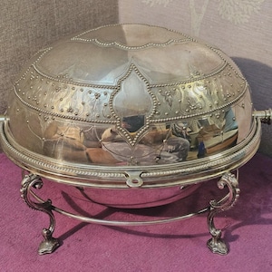 Puede incluir: Fuente de servir plateada y ornamentada, con tapa abovedada y detalles decorativos. La fuente se apoya en un soporte a juego con patas curvas. La tapa presenta intrincados patrones.
