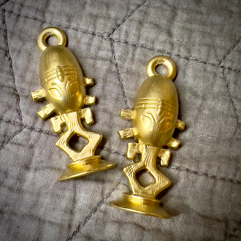 Puede incluir: Dos adornos de metal de color dorado con un dise&ntilde;o de rostro humano estilizado. Los adornos tienen forma de huevo con una base que se asemeja a una figura humana estilizada.