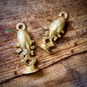Puede incluir: Dos amuletos de metal de color dorado en forma de figuras humanas estilizadas. Cada amuleto tiene un bucle en la parte superior para sujetarlo a un collar o pulsera.