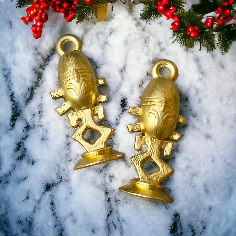 Puede incluir: Dos adornos decorativos de color dorado con dise&ntilde;os intrincados y un patr&oacute;n con forma de cara. Los adornos est&aacute;n sobre una superficie blanca cubierta de nieve.