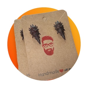 May include: Dark brown insect-shaped stud earrings on a brown card with a red illustration of a man with glasses and beard. The card reads "Handmade with love". The earrings are displayed on a bright orange and yellow background.