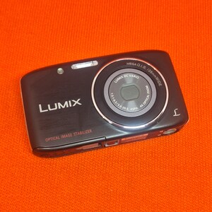 Könnte beinhalten: Schwarze Lumix Digitalkamera mit silbernem Objektiv und dem Text "LUMIX DC VARIO" und "OPTICAL IMAGE STABILIZER". Die Kamera hat einen 4-fach optischen Zoom und ein 28-mm-Weitwinkelobjektiv. Auf einer orangefarbenen Oberfläche.