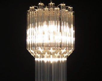 May include: A clear glass chandelier with a geometric design hangs from a chain against a black background. The chandelier is lit and the light shines through the glass.