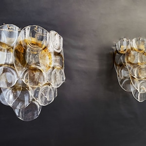 May include: Two clear glass wall sconces with a gold finish. The sconces are made up of multiple overlapping circular shapes.