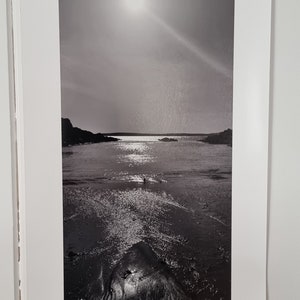 May include: A black and white photograph of a beach scene with a bright sun shining in the sky. The water is calm and reflects the sun's rays. The beach is sandy and there are rocks in the distance.