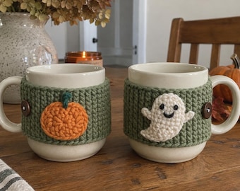 May include: Two cream-colored ceramic mugs, each wrapped in a textured olive green crochet cozy. One cozy features a small orange pumpkin appliqué, while the other displays a white ghost appliqué with a black face. Both are secured with brown buttons.