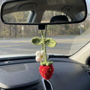 May include: A crocheted car charm in the shape of a strawberry, with green leaves and a white flower. The red strawberry has yellow detailing. The charm is suspended from a car's rearview mirror.