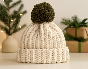 May include: A cream-colored, ribbed knit hat with a large olive green pom-pom on top. The hat has a folded brim and is likely made of a soft, warm material. The hat is a winter accessory.