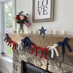 May include: A decorative garland featuring a series of interconnected bows in red, white, and blue yarn. The bows are strung together on a white cord, creating a repeating pattern of colours that hangs horizontally across a wooden fireplace mantel.