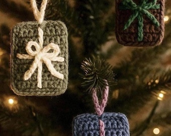 May include: Crocheted Christmas ornaments in green, brown, and blue, each shaped like a gift box. The ornaments feature a contrasting bow and hanging loop. The background is a blurred image of a Christmas tree with lights.