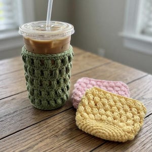 Op de afbeelding: Een doorzichtige plastic beker gevuld met een lichtbruine ijsdrank en een wit rietje, voorzien van een saliegroene gehaakte hoes met een textuur van nopjessteken. Twee extra gehaakte hoezen in pastelgeel en roze liggen ernaast op de ondergrond.