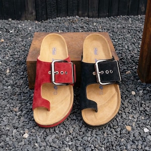 May include: A pair of open-toe sandals, one red and one black, each with a large buckle. The sandals rest on a wooden block against a backdrop of dark wood and gravel. The sandals have a toe loop design.