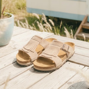 May include: A pair of beige sandals with fringe detailing and adjustable buckles, resting on a wooden surface. The sandals have a cork footbed and are set against a backdrop of potted plants and a vintage camper.