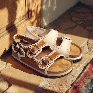 May include: A pair of leather sandals with adjustable buckles. One sandal is brown with white stitching, the other is white with white stitching. Both have cork footbeds and are displayed on a stone surface.
