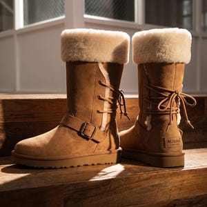 May include: A pair of tan suede mid-calf boots featuring a thick cream-colored shearling lining, a fold-over cuff, and a lace-up detail along the side. Each boot includes a decorative strap with a bronze-toned metal buckle and an AU&MU label.