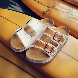 May include: Two pairs of sandals with cork footbeds and leather straps rest on yellow surfboards. One pair is white with a single strap, and the other is brown with two straps. Both have metal buckles and decorative stitching.