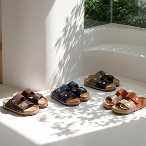 May include: A collection of open-toe sandals in various colors, including brown, navy blue, olive green, and tan. Each pair features adjustable straps and cork footbeds. The sandals are displayed on a white surface with natural light.