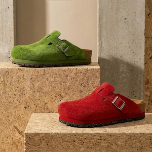 May include: Two suede clogs, one green and one red, are displayed on a textured surface. The green clog is on a higher level. Both clogs have a buckle closure and a cork footbed. The shoes are a casual style.