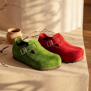 May include: A pair of suede shoes, one green and one red, with a strap and buckle closure. The shoes are displayed on a white surface, with a neutral background. The shoes have a textured appearance.