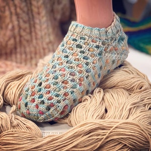 May include: A pair of colorful knitted socks with a lacy pattern. The socks are made with a variety of colors, including blue, green, pink, and orange. The socks are sitting on a pile of beige yarn.