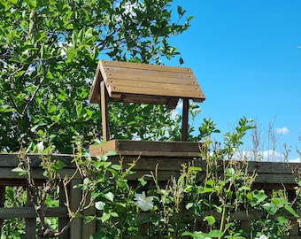 May include: A wooden bird feeder with a brown stain finish. The feeder has a flat roof and a tray for birdseed. The feeder is mounted on a wooden fence post.