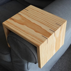 Puede incluir: Una bandeja de madera para el reposabrazos de un sofá o sillón. La bandeja está hecha de madera clara con un acabado natural y tiene una forma rectangular. Está diseñada para encajar en el reposabrazos de una silla y proporcionar una superficie plana para sostener bebidas, refrigerios u otros artículos.
