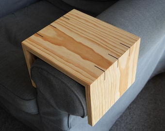 May include: A wooden armrest tray for a couch or armchair. The tray is made of light wood with a natural finish and has a rectangular shape. It is designed to fit over the armrest of a chair and provide a flat surface for holding drinks, snacks, or other items.