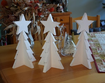 May include: Four white wooden Christmas trees with star toppers. The trees are arranged on a wooden table with a patterned tablecloth.