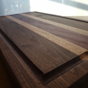 May include: A large, rectangular wooden cutting board with a raised edge. The board is made of walnut, maple, and purpleheart wood. The wood grain is visible and the board has a natural finish.